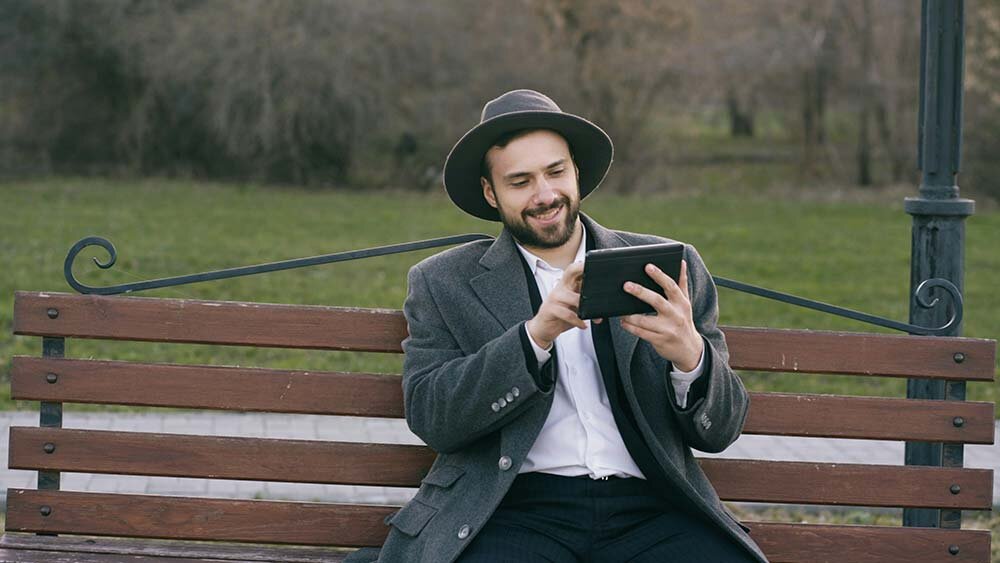 A man wearing a hat and seated on a park bench using his tablet to apply for a direct loan.