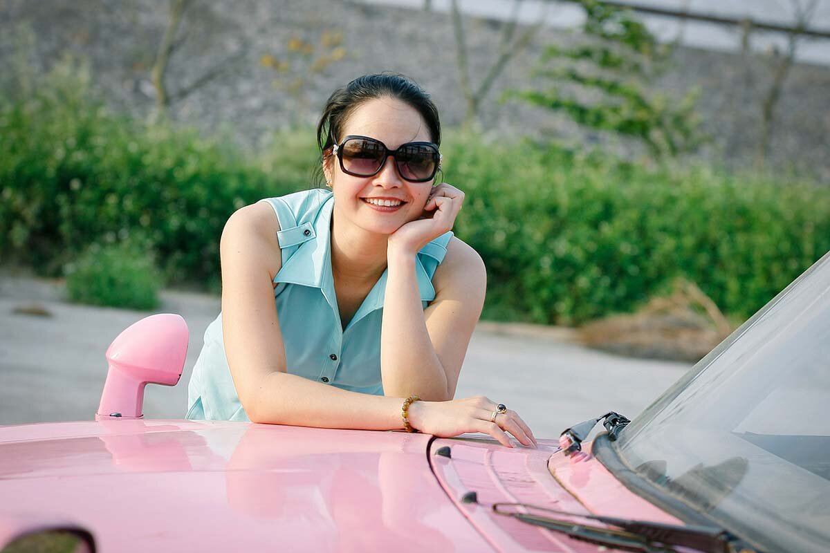 A happy young woman leaning forward against a parked pink car.