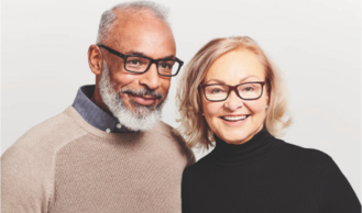 An older man with a white beard and glasses stands next to an older woman with glasses, both smiling, against a light gray background.