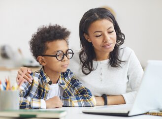 A woman and a child are sitting at a table, looking at a laptop screen. The woman has her arm around the child. The table has pencils and a notebook