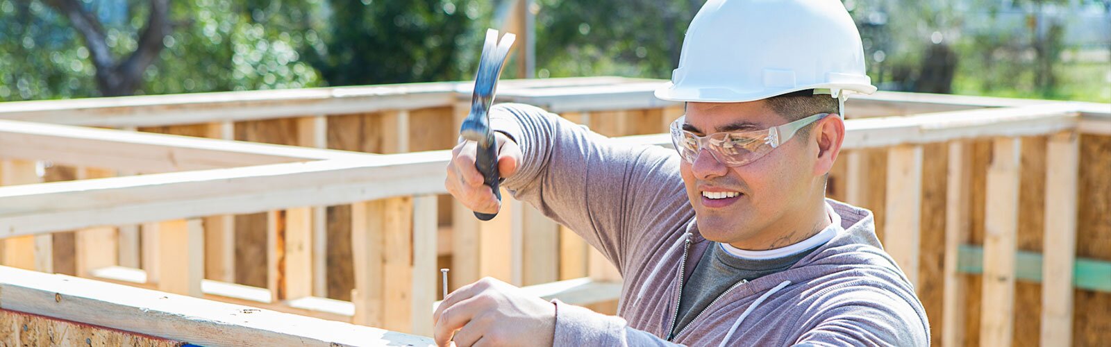 A contractor wearing a hard hat and safety glasses hammers nails into framing on a house under construction.