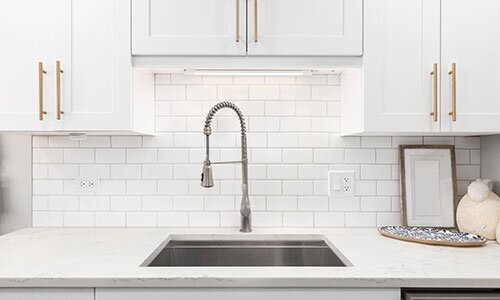 New kitchen sink and faucet with bright lighting above, white countertops and cabinets with gold hardware.