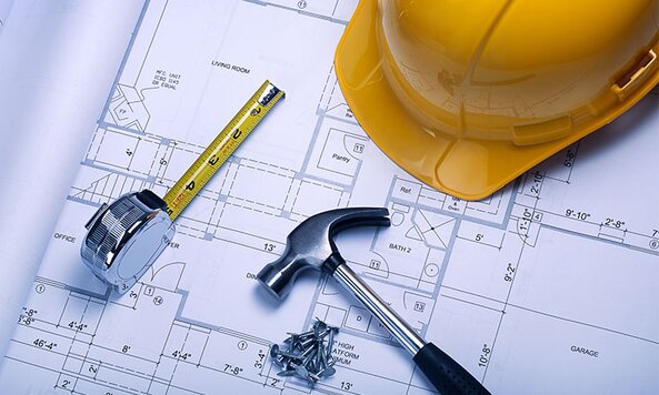 A yellow hard hat sits on top of blueprints with a tape measure, a hammer, and nails. 