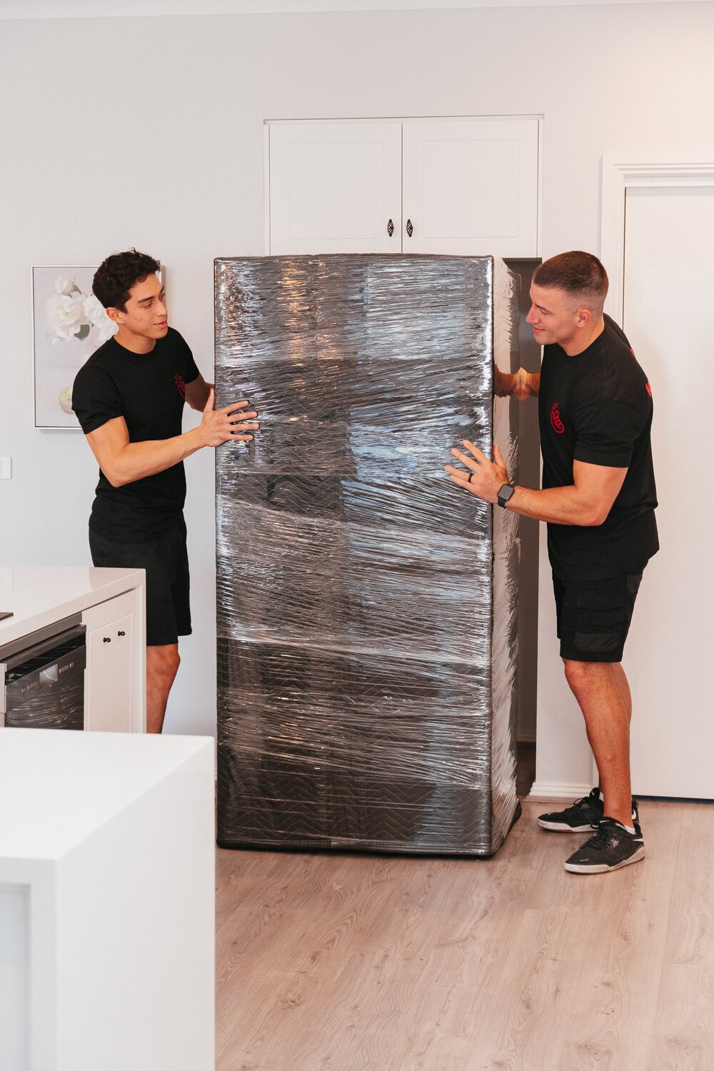 Instamove removalists wrapping a stainless steel fridge with a protective moving blanket in a modern kitchen before an interstate move