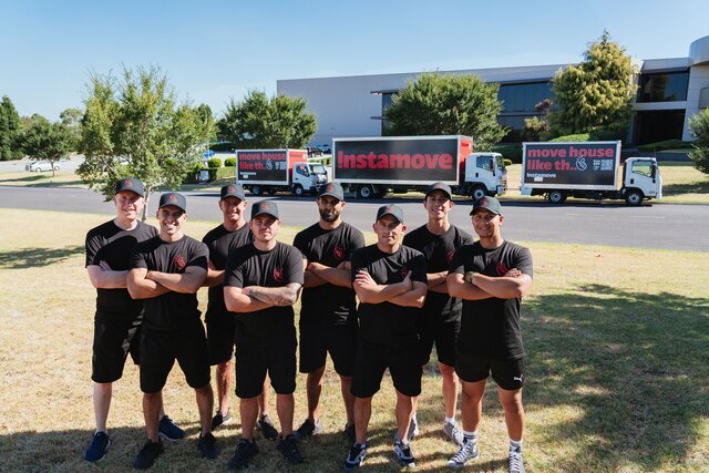 Instamove team in branded uniforms posing in front of three moving trucks on a suburban lawn