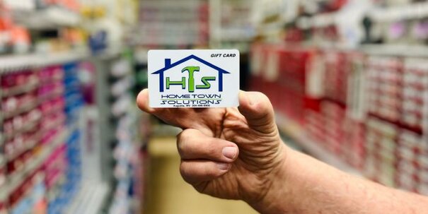 A photo of a hand holding the HomeTown Solutions gift card with the hardware aisle in the background.
