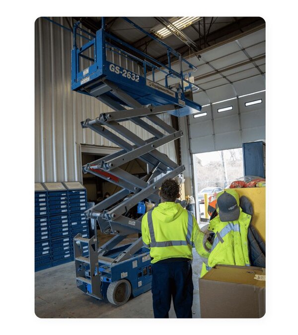 A Genie GS-2632 scissor lift raised in the air with a service technician watching from below