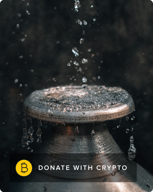 A woman smiles and faces the camera while washing her clothes using an outdoor faucet.