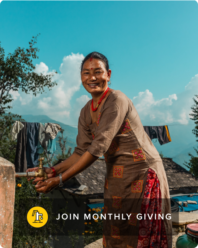 A woman smiles and faces the camera while washing her clothes using an outdoor faucet.