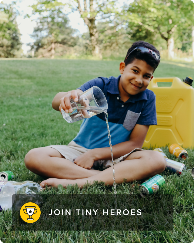 A young boy sits down cross-legged and pours a glass of water into a grassy patch.