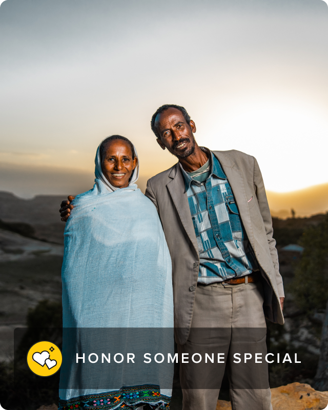 A man and woman stand together, smiling directly into the camera against a backdrop of mountains.