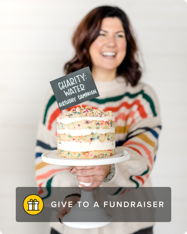 A woman with a striped multi-color sweater smiles while holding a colorful cake with a decoration sign that says, "charity: water".