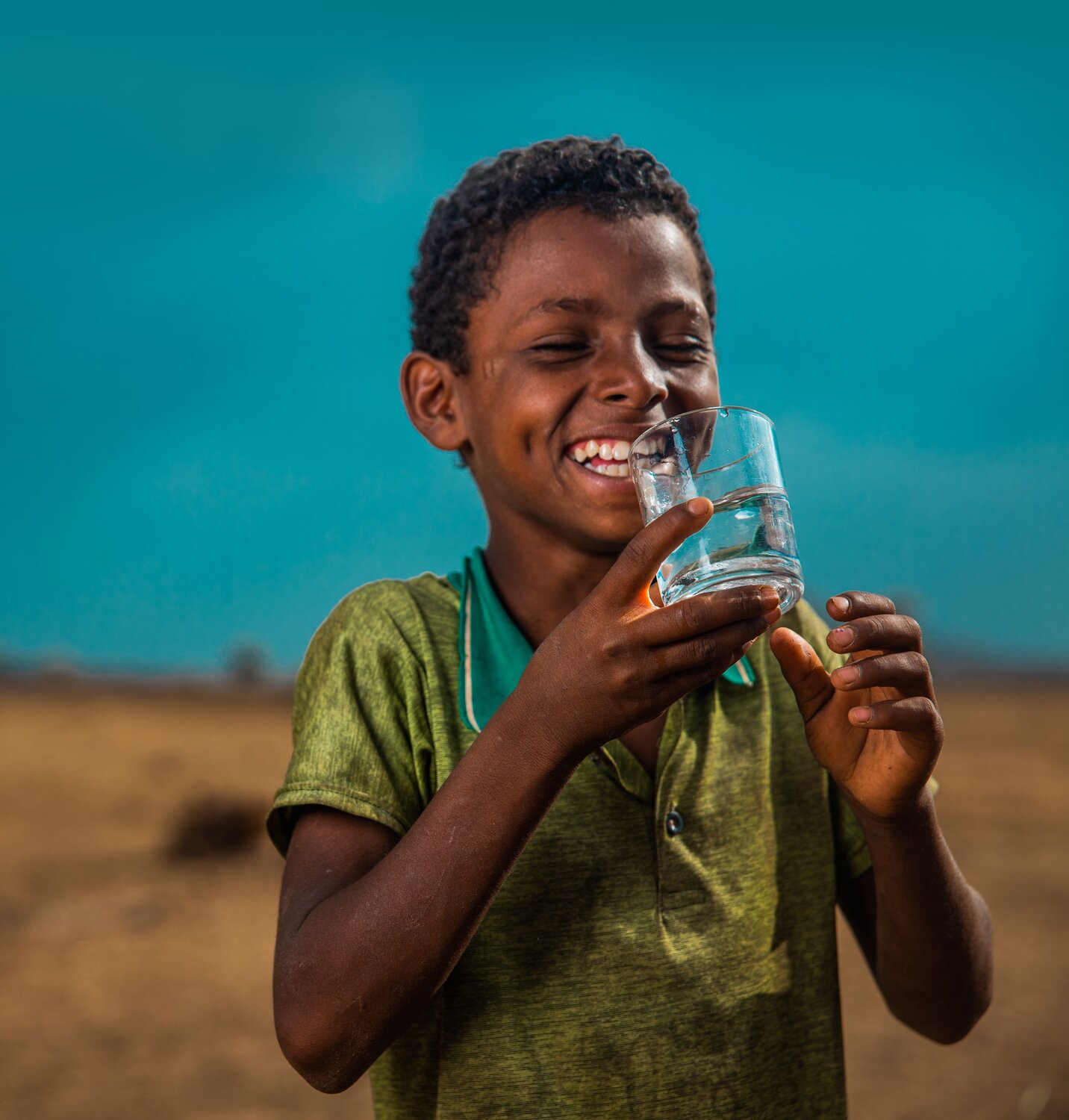 Young boy with a green short-sleeve shirt holds a glass of water and smiles.