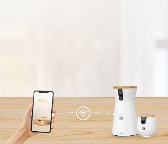 Two Furbo pet cameras sit on a wooden table, connected via a Wi-Fi symbol to a smartphone held in a hand showing the Furbo app.