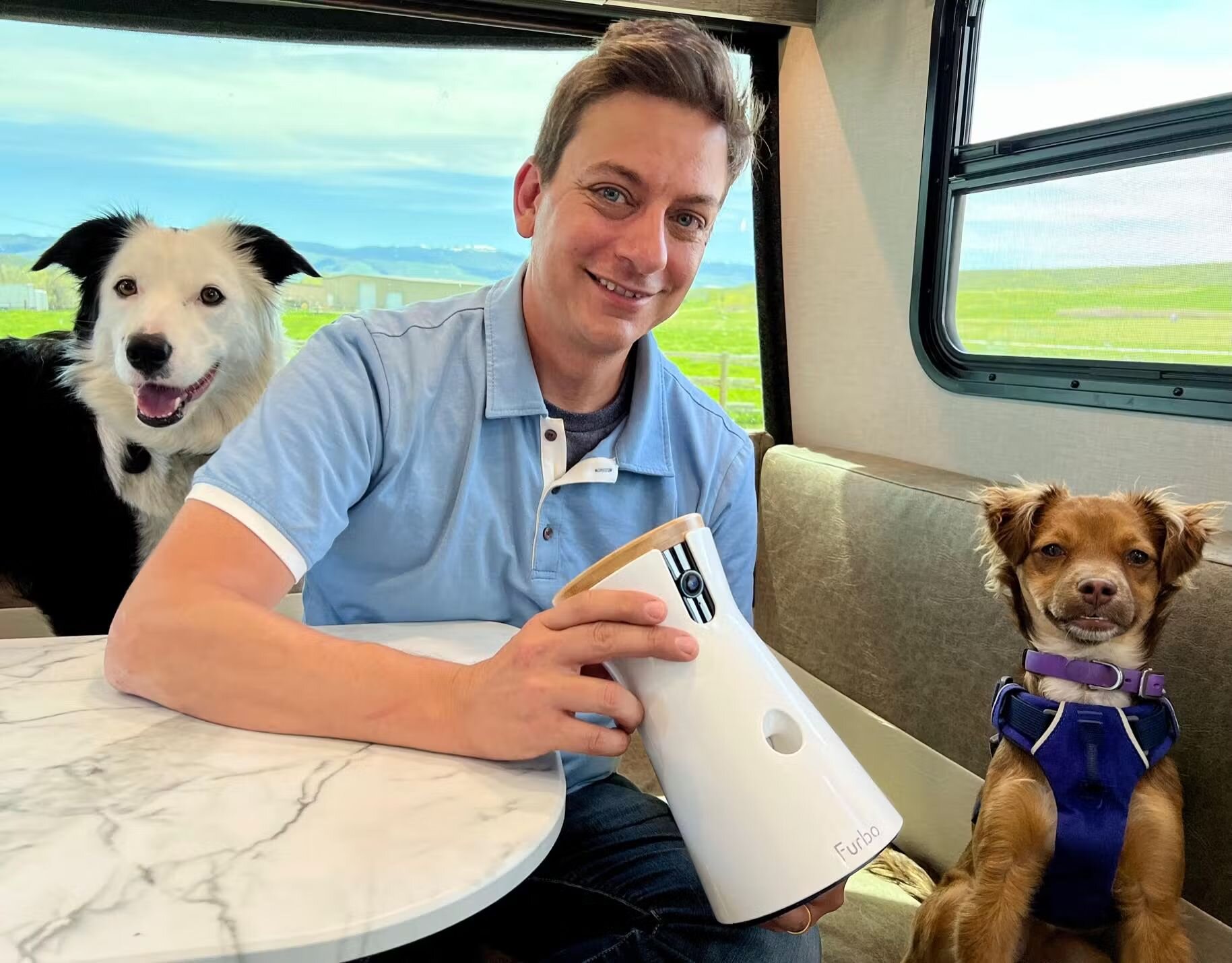Smiling man holds a Furbo Dog Camera in an RV, with two happy dogs sitting beside him