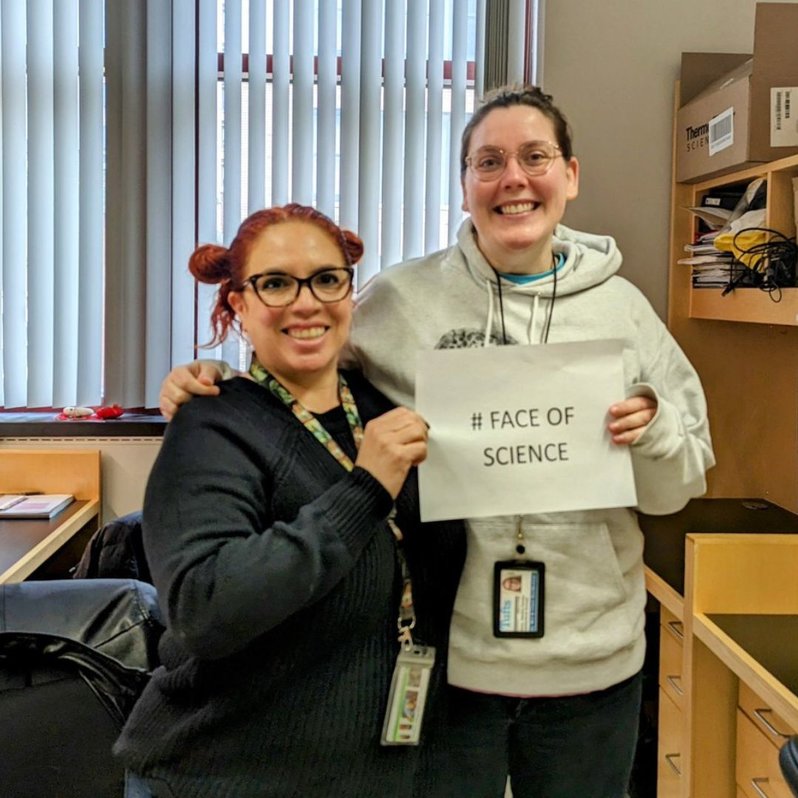 Sandra Sanchez PhD with a lab mate smiling proudly and holding a #FaceOfScience sign