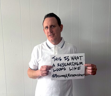 Zak Peet holds a sign that says “This is what a researcher looks like #BecomeAResearcher"