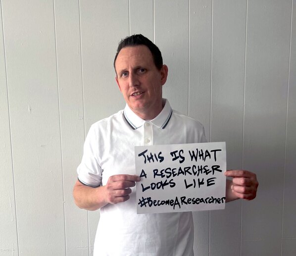 Zak Peet holds a sign that says “This is what a researcher looks like. “#BecomeAResearcher”
