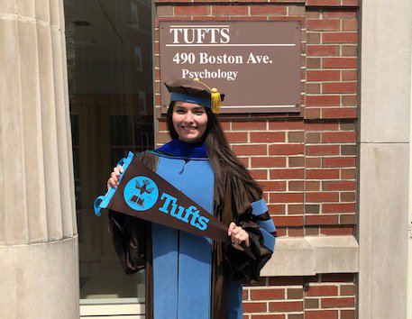 Cecilia Hinojosa in front of the Psychology building at Tufts University. She is in her graduation cap and gown proudly holding a Tufts flag.