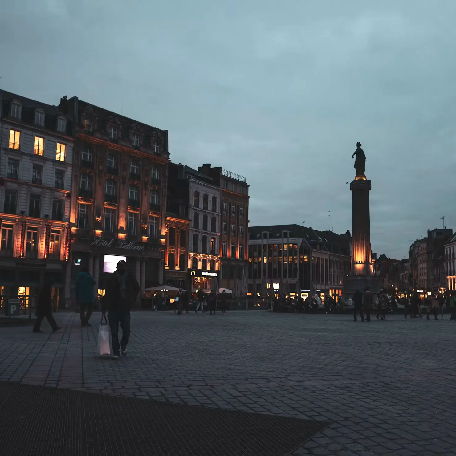 Illuminated statue and column in a Lille city square at dusk