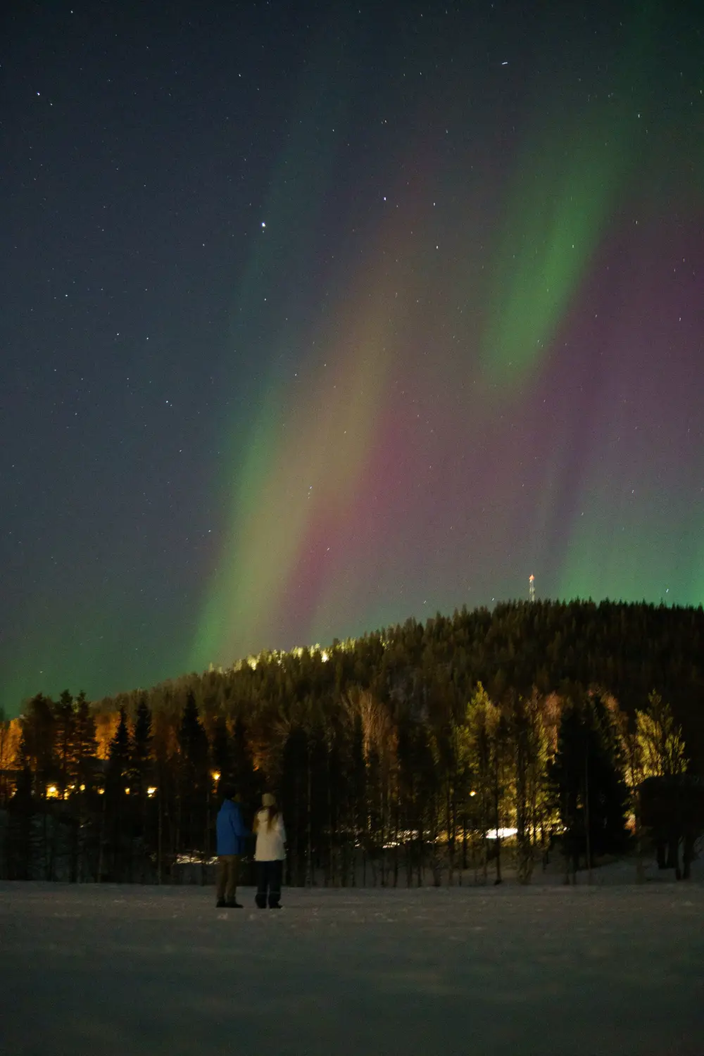 Two people standing on a snowy field beneath colorful northern lights over a forested hillside at night.