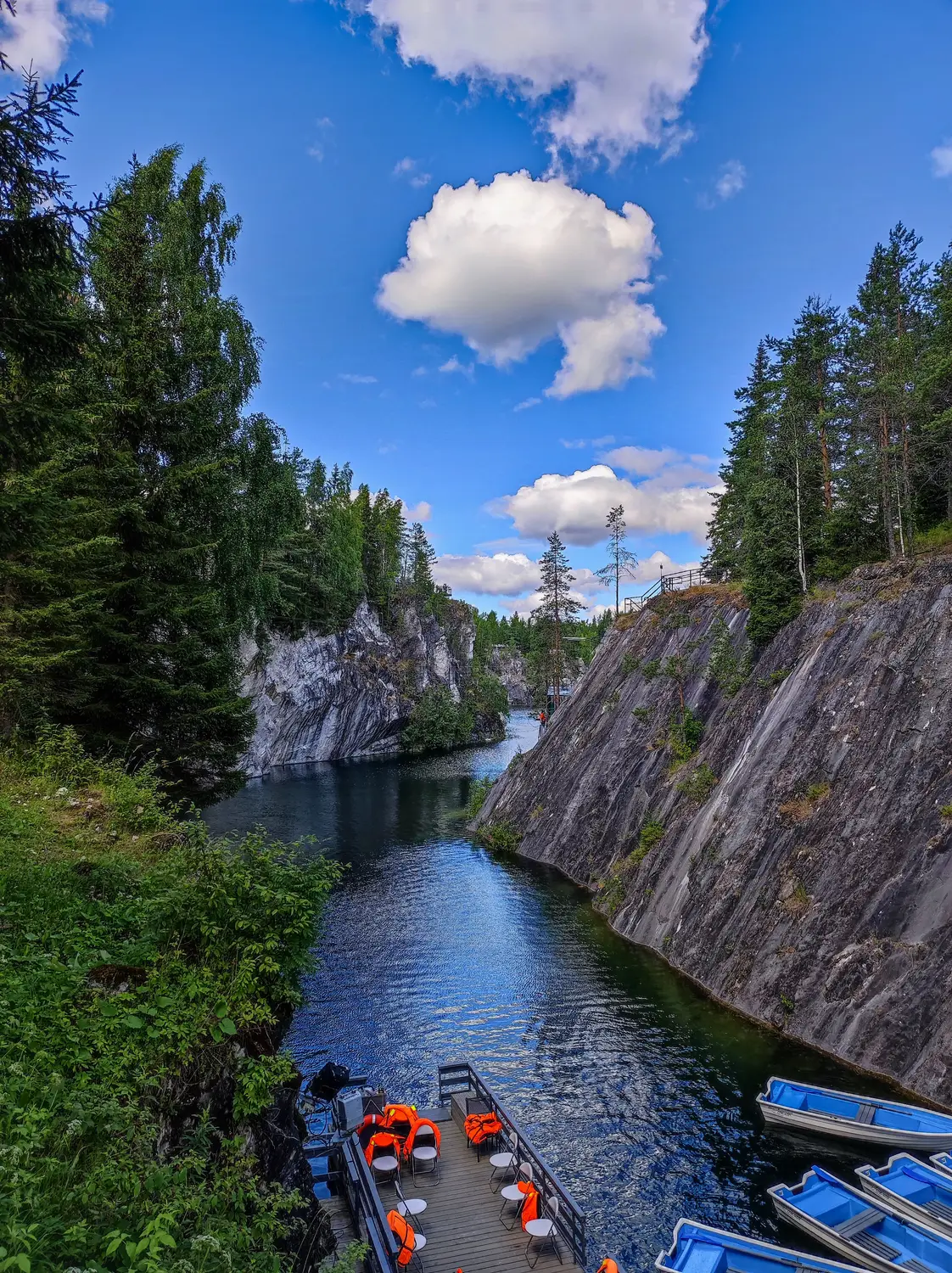 Narrow river flowing through a rocky forest gorge, with small blue boats and a dock beneath a bright blue sky with clouds.