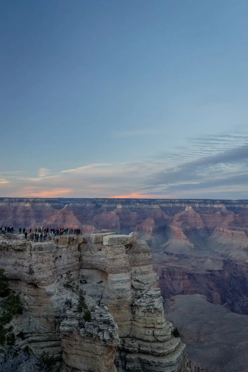 Visitors gathered on a rocky overlook at the Grand Canyon at dusk, with layered canyon walls fading into the twilight sky.