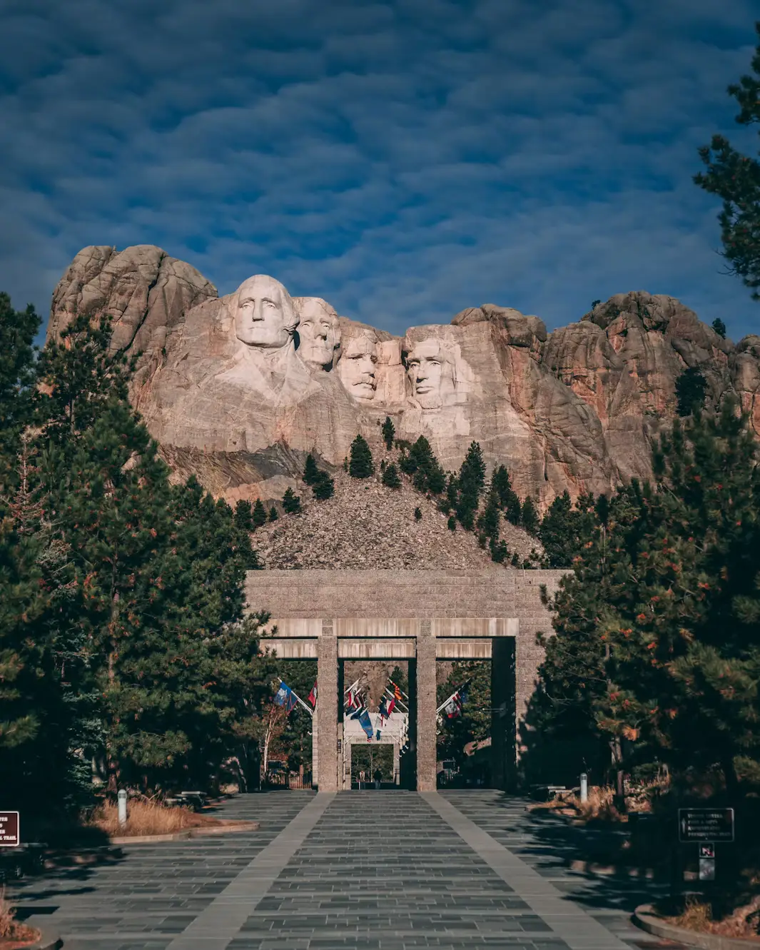 The carved presidential faces of Mount Rushmore rising above the flag-lined Avenue of Flags under a cloudy blue sky.