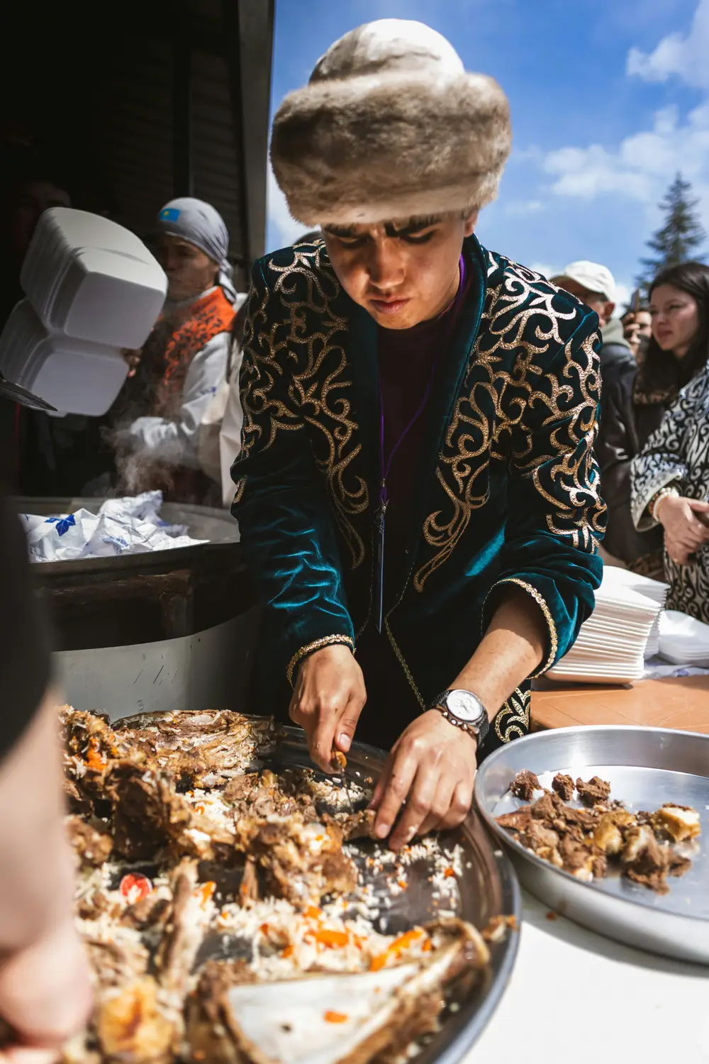 A focused young man in a teal gold-embroidered jacket and fur hat serving traditional plov rice from a large communal pan outdoors