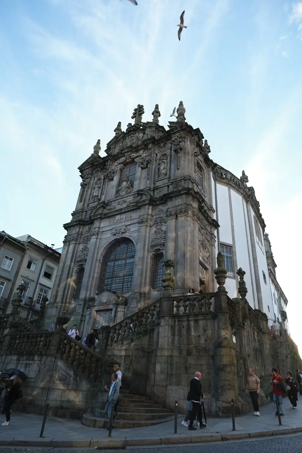 An ornate baroque church with grand stone steps, people walking outside, and birds flying overhead.