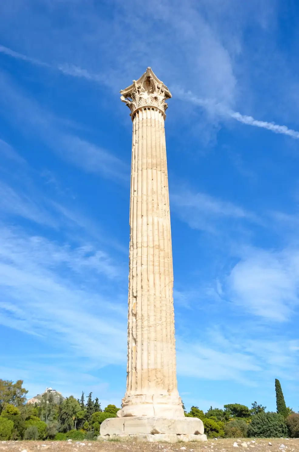 A tall, standalone ancient Corinthian column with a decorative capital, set against a blue sky with trees below.