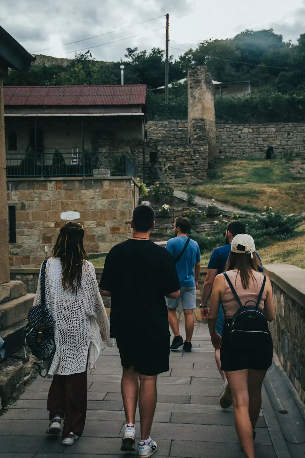 Group of people walking through a stone-paved village street with old buildings and ruins under a cloudy sky.
