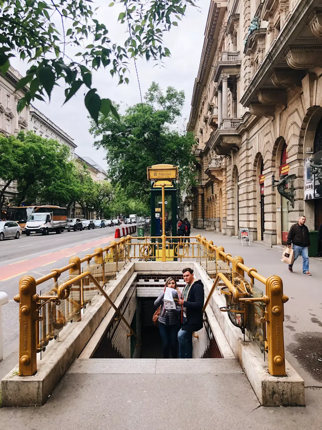 Historic subway entrance with ornate yellow railings on a tree-lined city street, with two people holding a map.