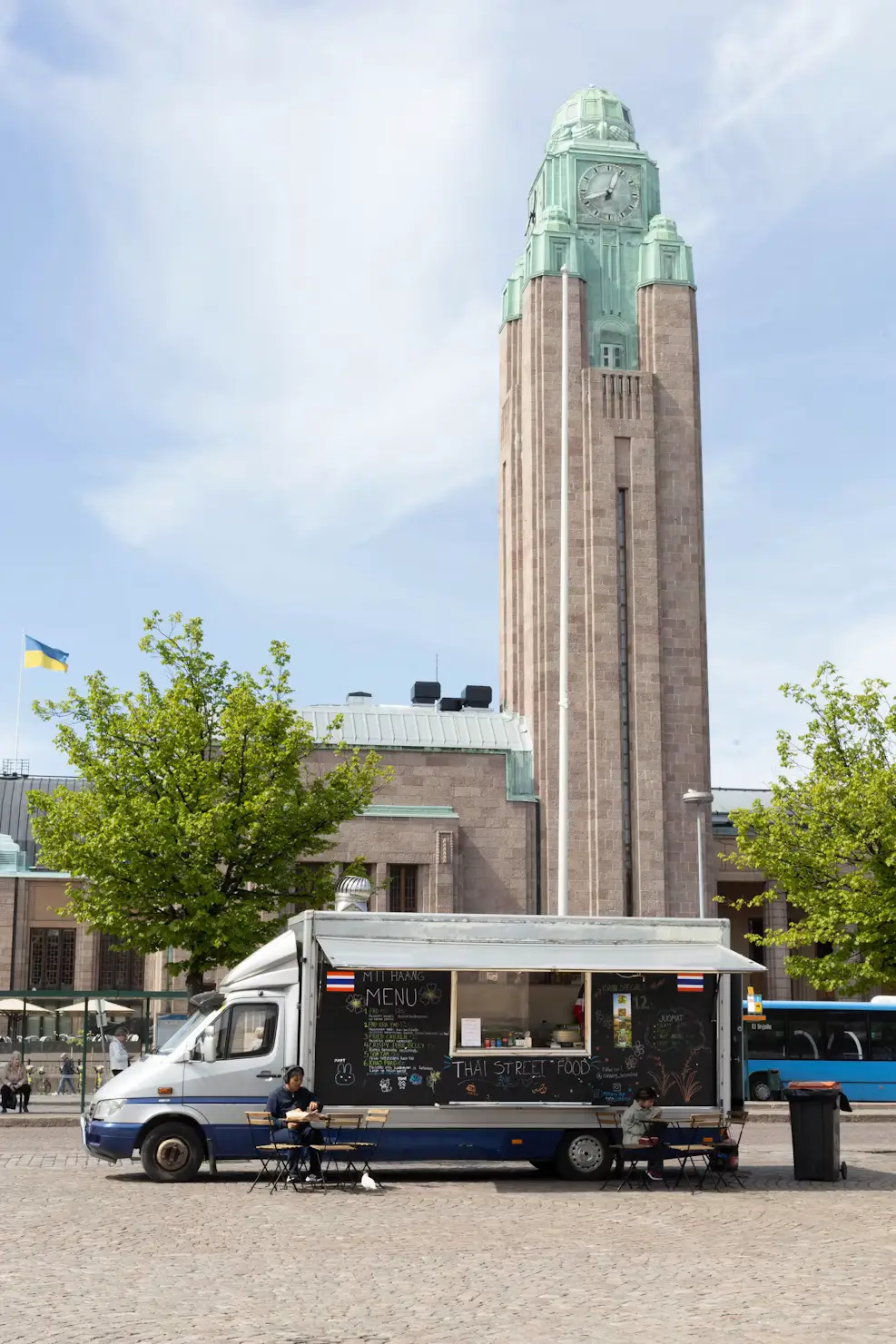 Thai street food truck parked in a cobblestone city square with outdoor seating, set in front of a tall stone clock tower and leafy trees under a clear sky.