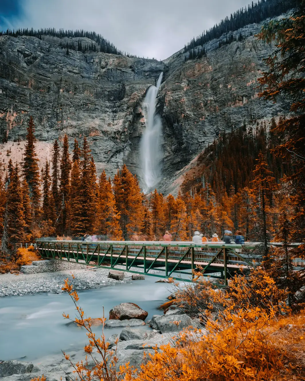 A tall waterfall cascading down a rocky cliff into a valley of orange autumn trees, with a footbridge crossing below.