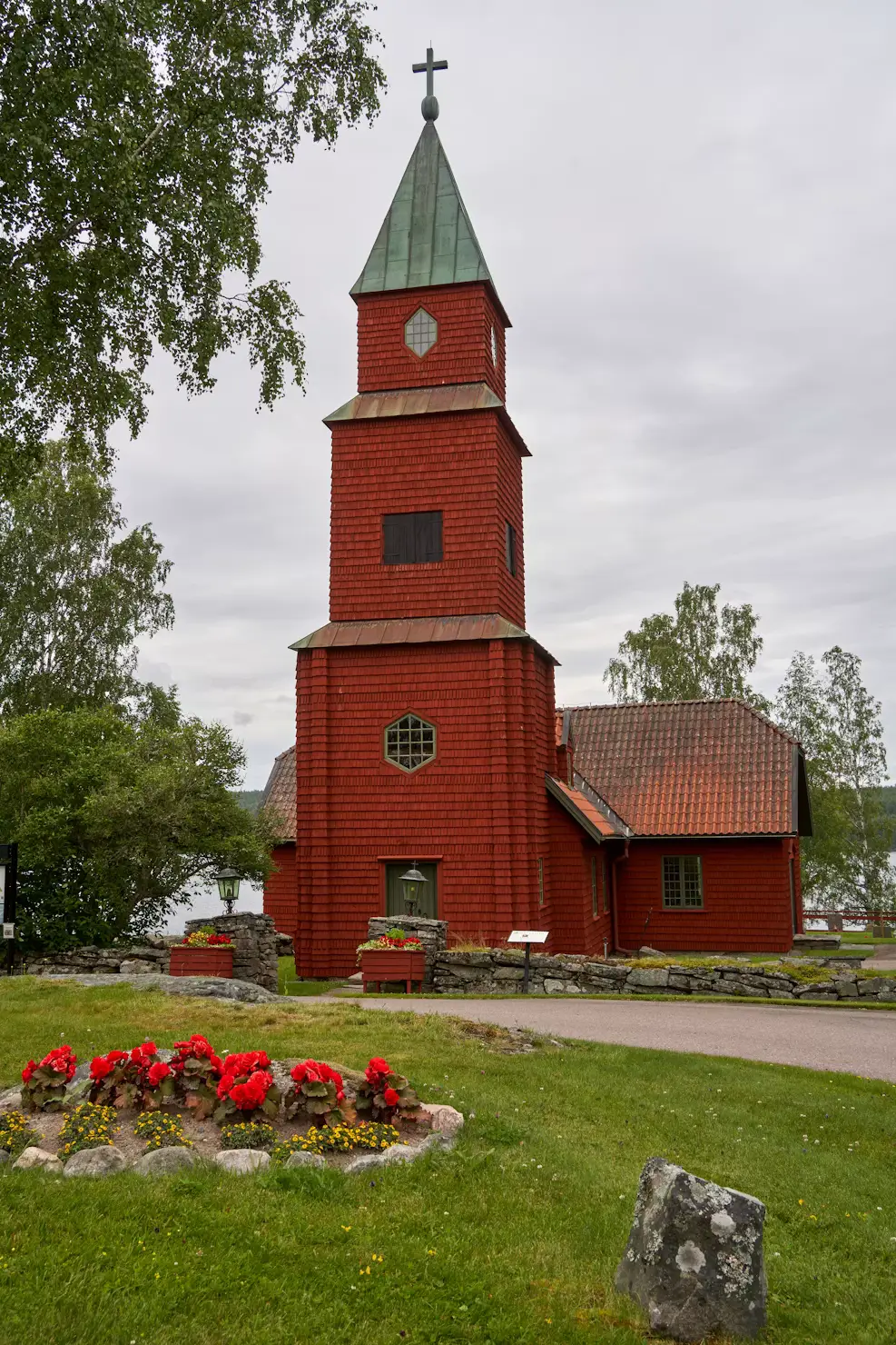 Small red wooden church with a tall bell tower, set on a lawn with flower beds.