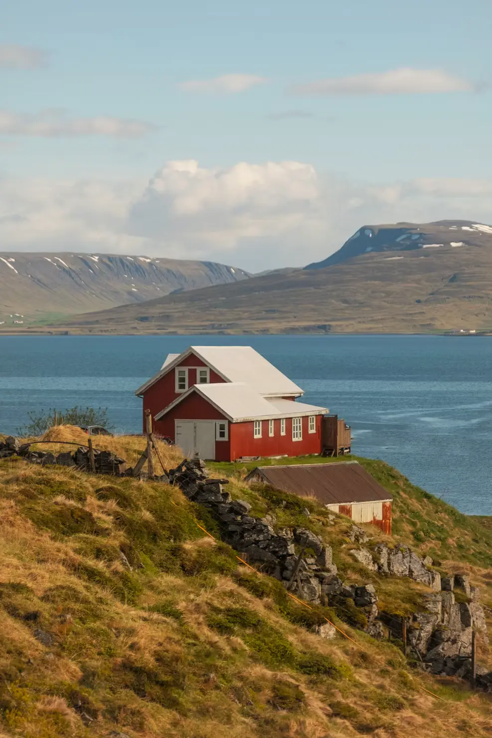 Red house on a grassy coastal hillside overlooking a fjord, with snow-capped mountains in the distance, Iceland.