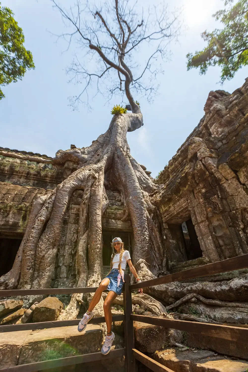 Female traveler posing in front of giant tree roots engulfing stone ruins at Ta Prohm temple, Angkor Archaeological Park, Siem Reap, Cambodia