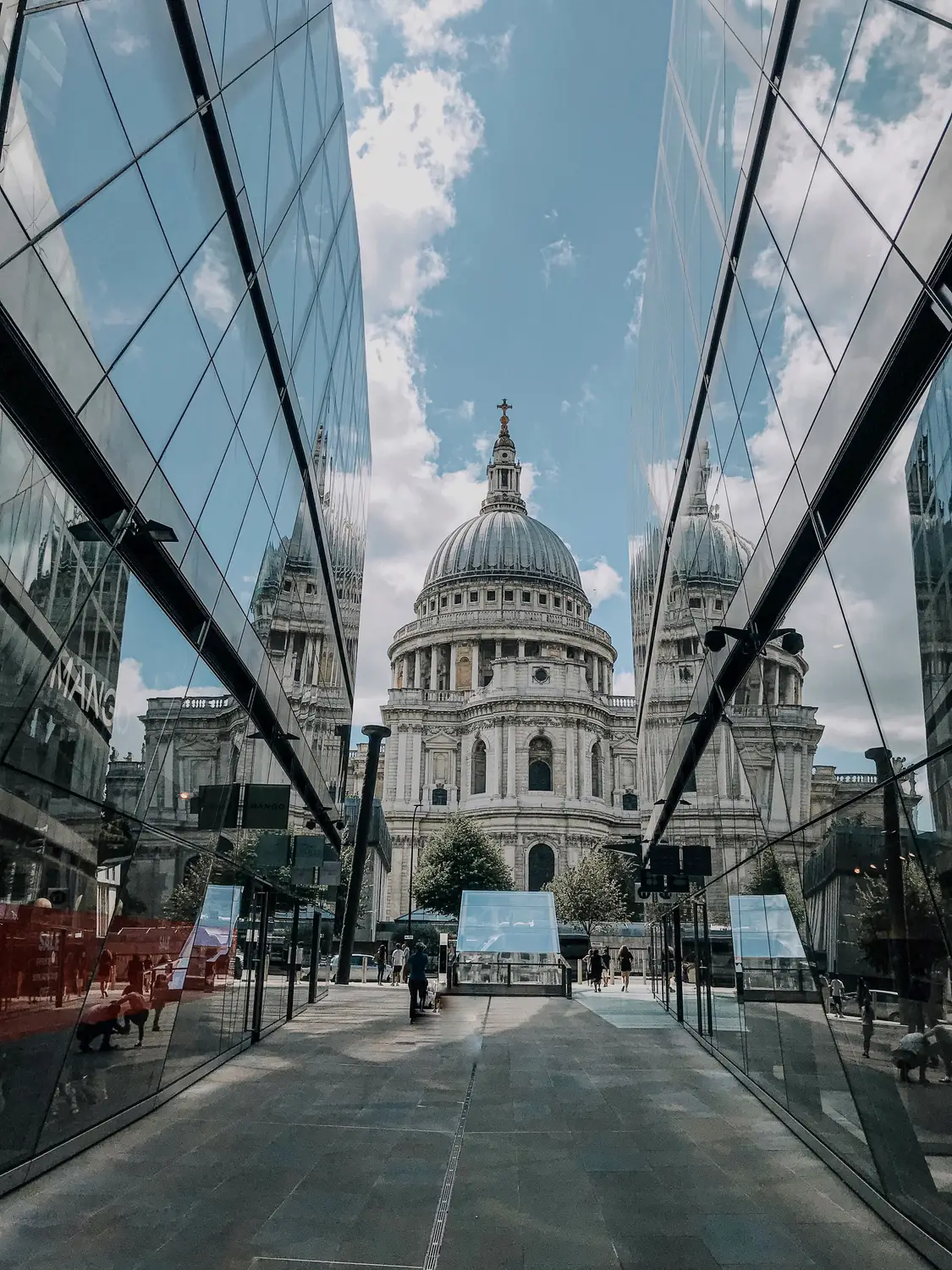 St Paul's Cathedral and its dome reflected in the angled glass facade of a modern building beside a sunlit street.