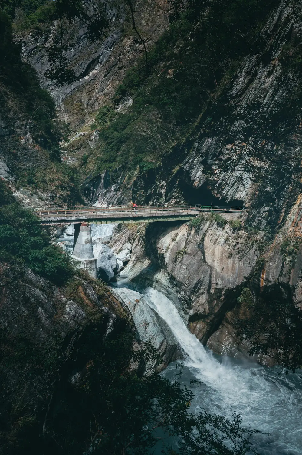 Person in red standing on a bridge over a waterfall in Taroko Gorge, surrounded by steep forested marble cliffs