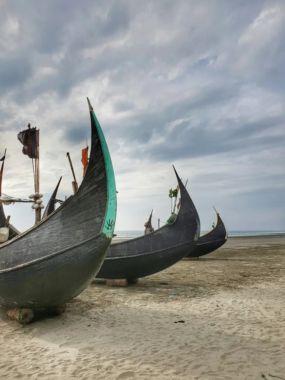 Traditional wooden Macher Dinghi fishing boats with curved bows resting on Cox's Bazar sandy beach under dramatic cloudy sky, Bangladesh