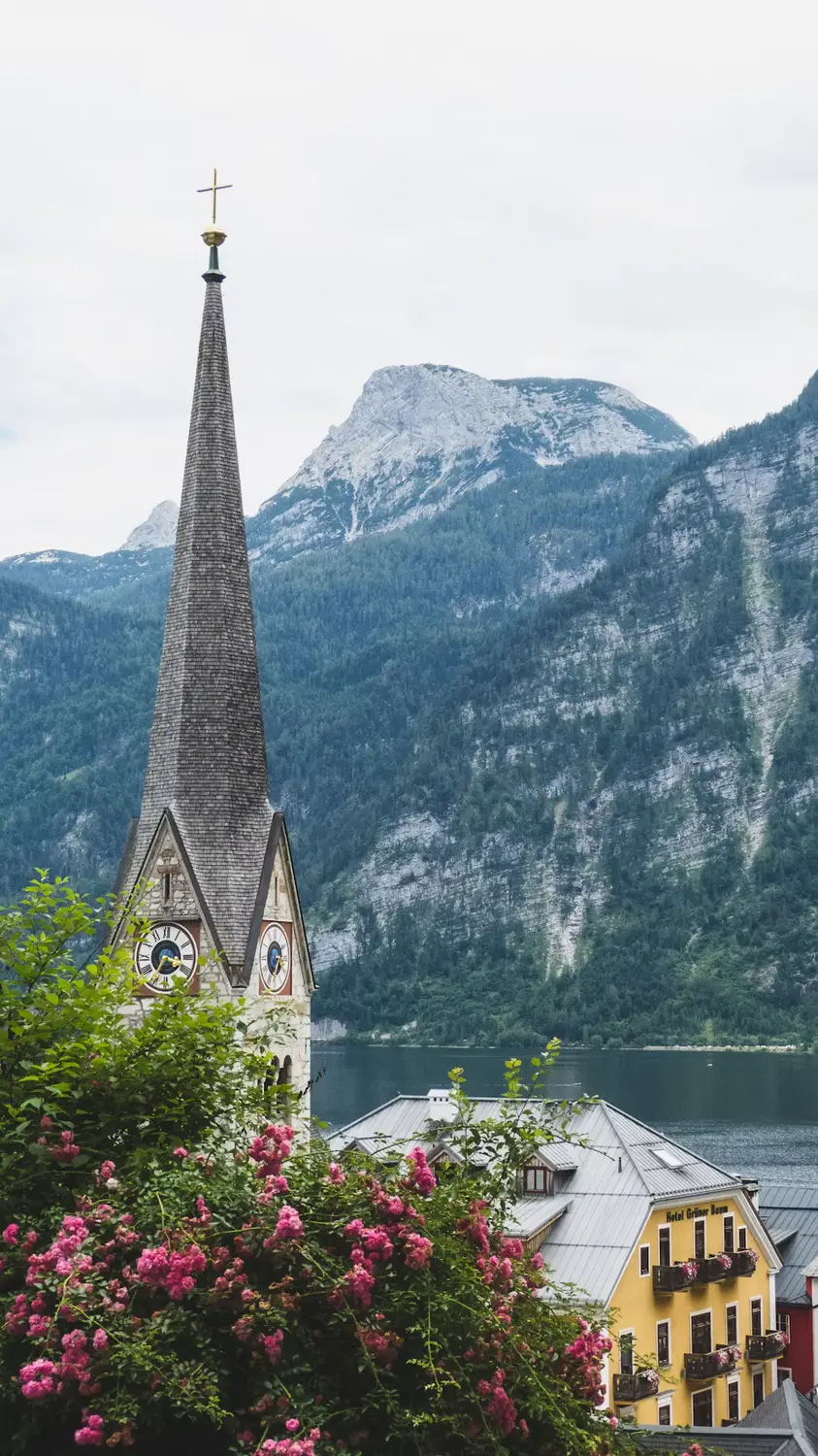 A stone church spire framed by pink flowers, overlooking a lakeside village and mountains in the background.