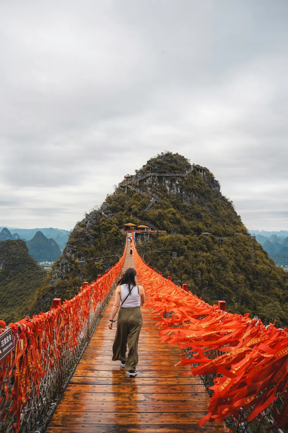 Woman walking on suspension bridge covered in red prayer ribbons leading to karst mountain peak in Guangxi China