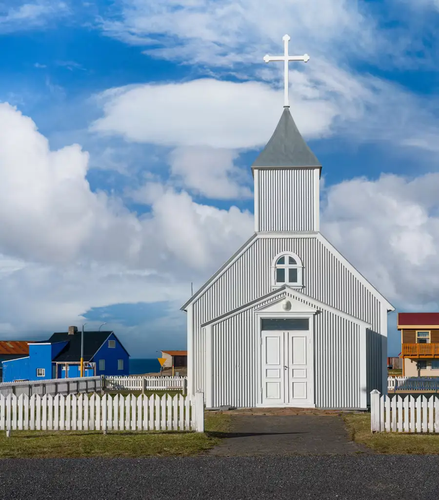 Small white church with a steep grey roof and cross, colorful houses visible behind a white picket fence, Iceland.