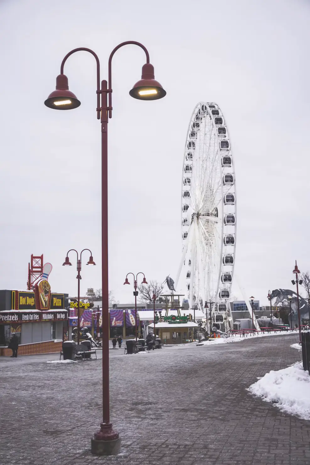 A snowy waterfront boardwalk with vintage lampposts and a Ferris wheel in the background under an overcast sky.
