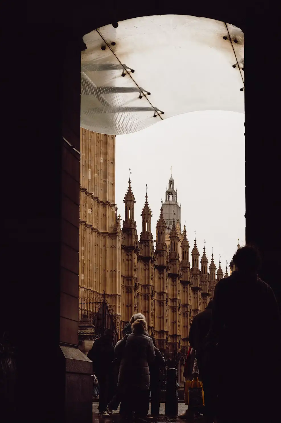 Silhouetted pedestrians framed by a dark archway, looking toward the gothic spires of the Houses of Parliament.