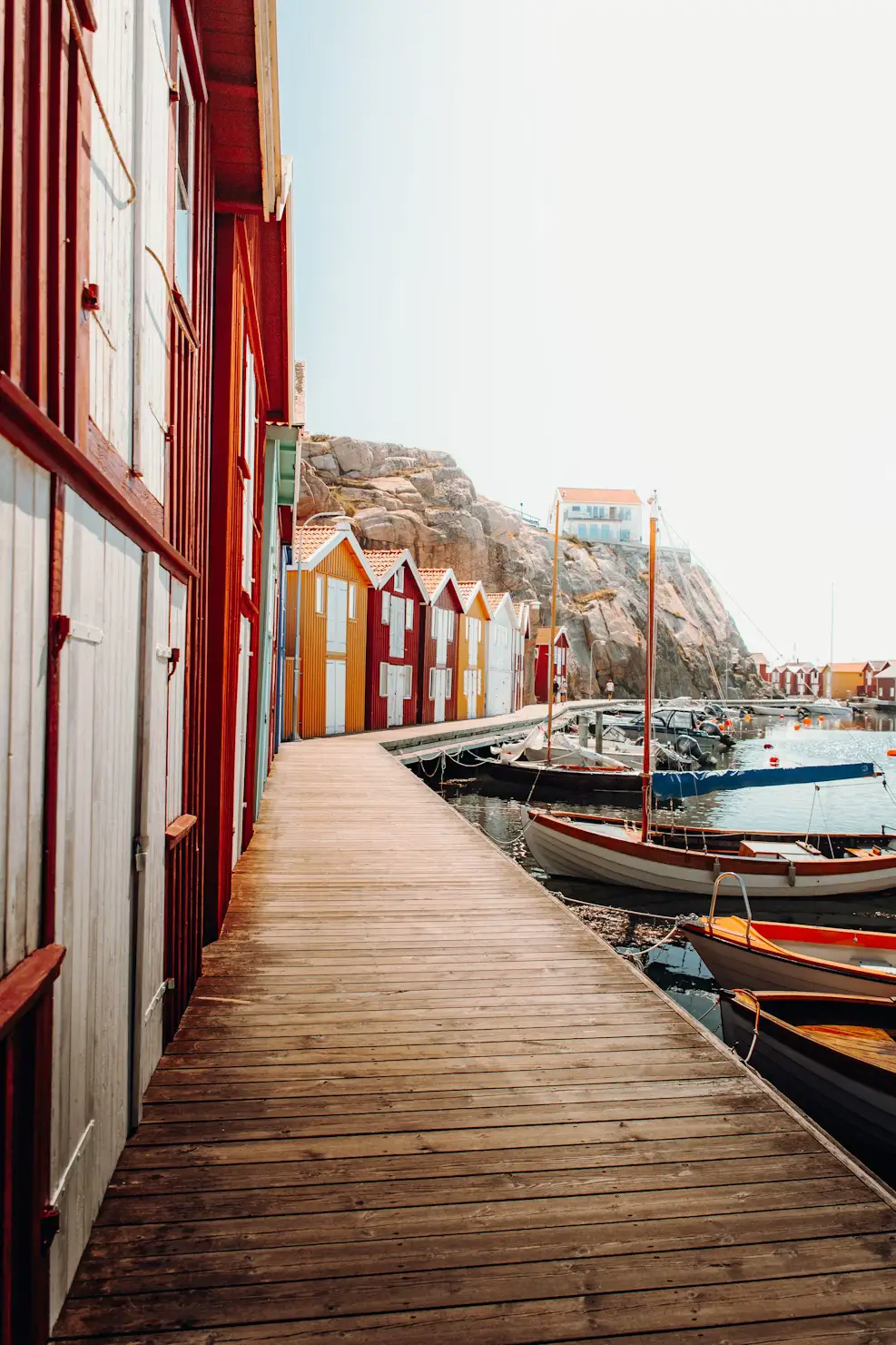 Wooden boardwalk beside red buildings leading to boats moored along the water, with a rocky hill in the background.