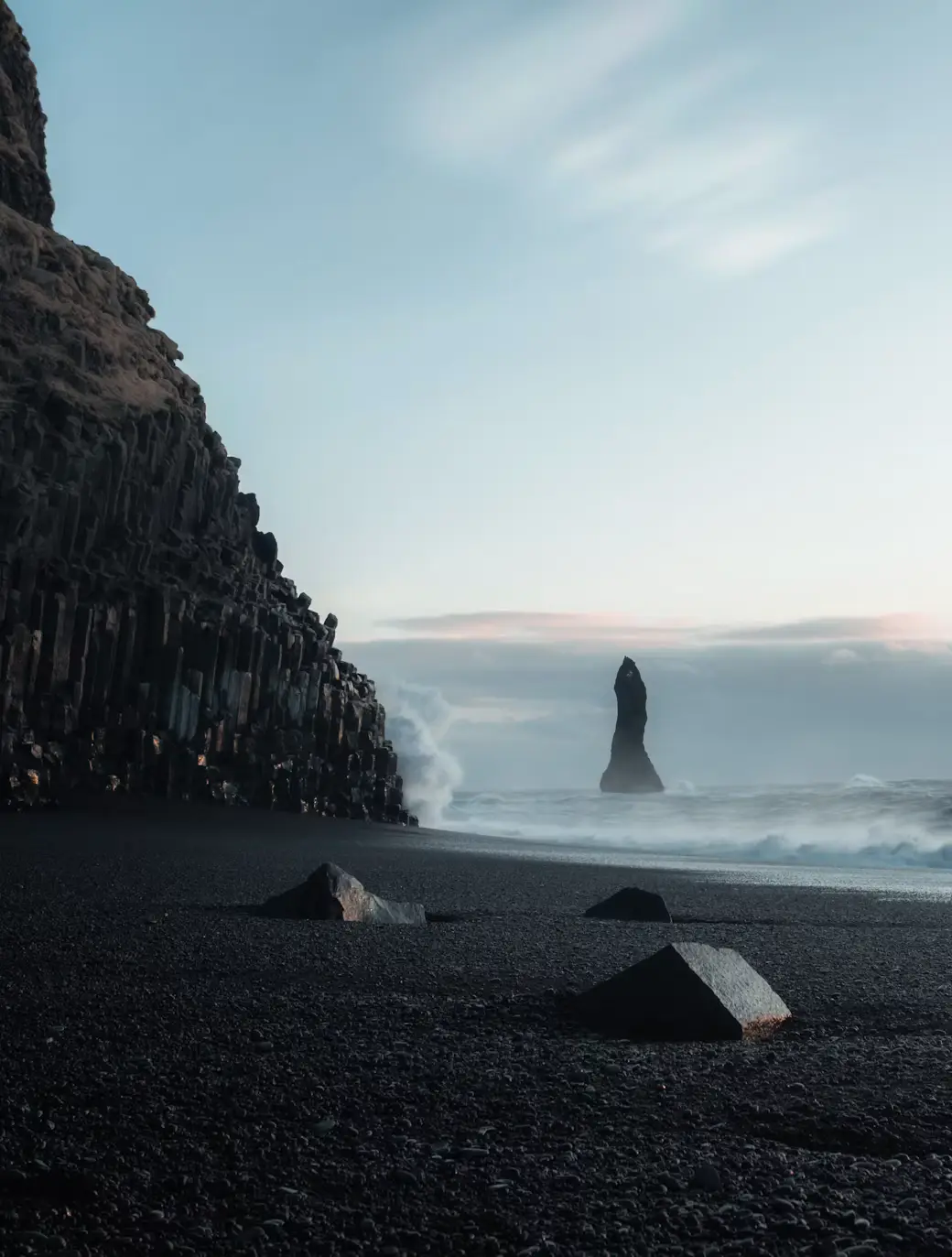 Black sand beach with a rock sea stack and cliffside, sea and soft sky in the background, Iceland.