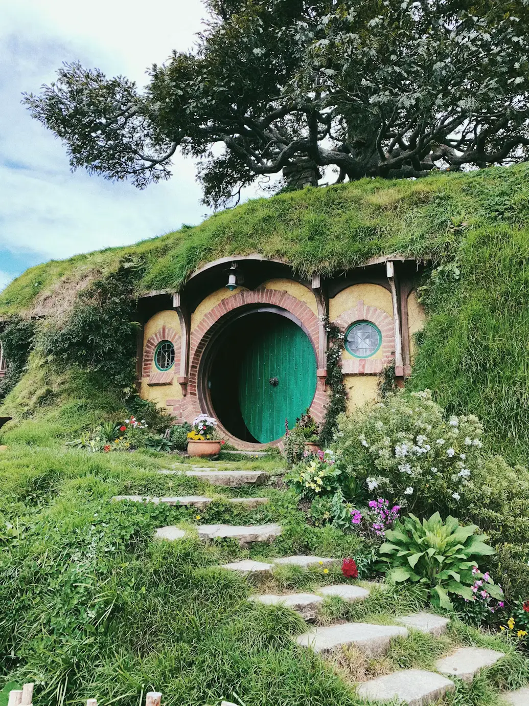 Hobbit-style grass-covered house with a round green door, stone path, and flowering garden.