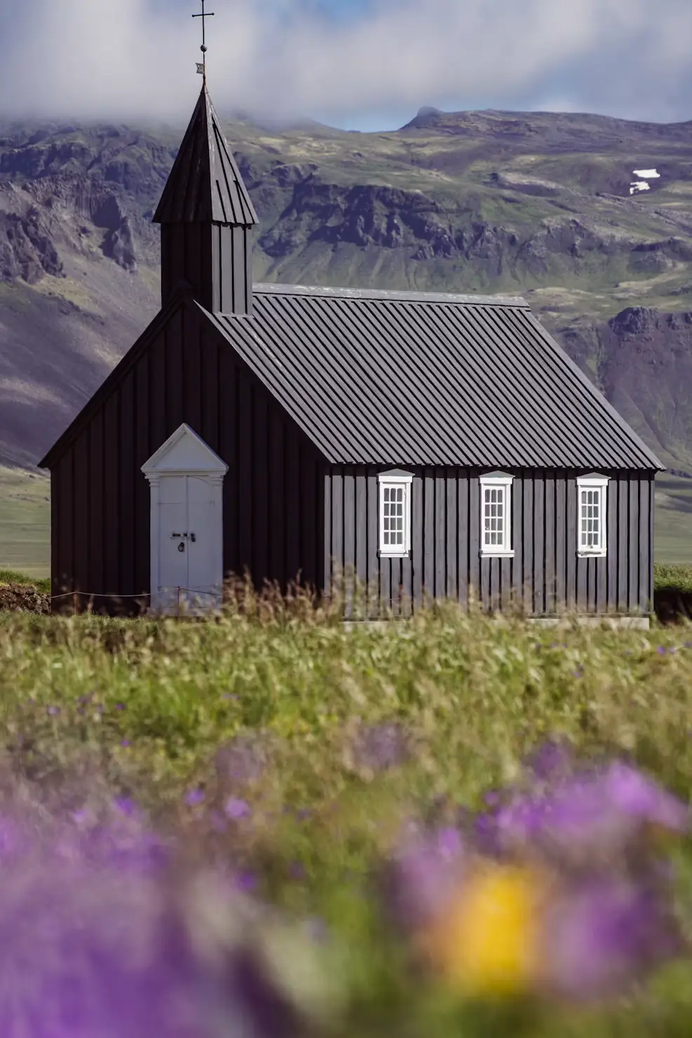 Black wooden church surrounded by purple wildflowers, with mountains rising behind, Iceland.